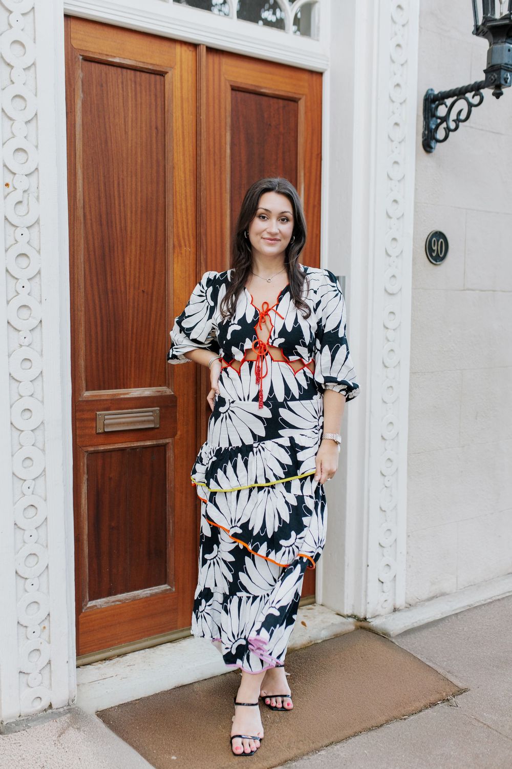 Woman in black and white floral maxi dress poses by wooden doors with ornate white trim.