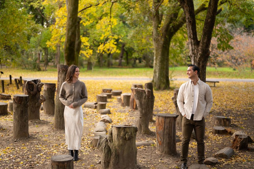 Couple smiling at each other among rustic tree stumps in a golden autumn park setting.