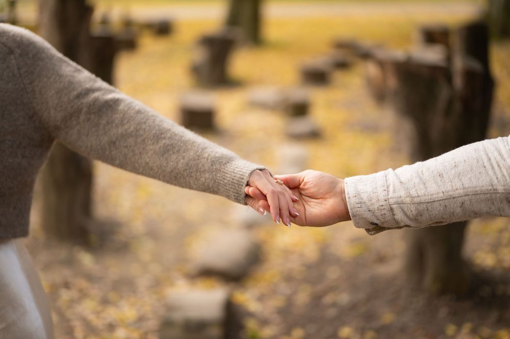 Close-up of couple holding hands gently among tree stumps with golden autumn leaves in background.
