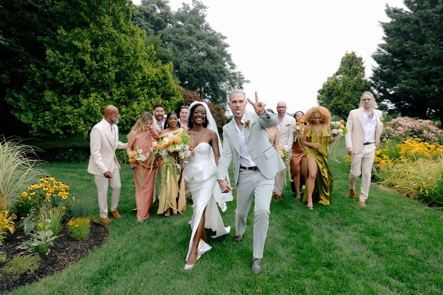 Wedding party celebrates on a lush garden lawn with colorful flowers and greenery in the background.