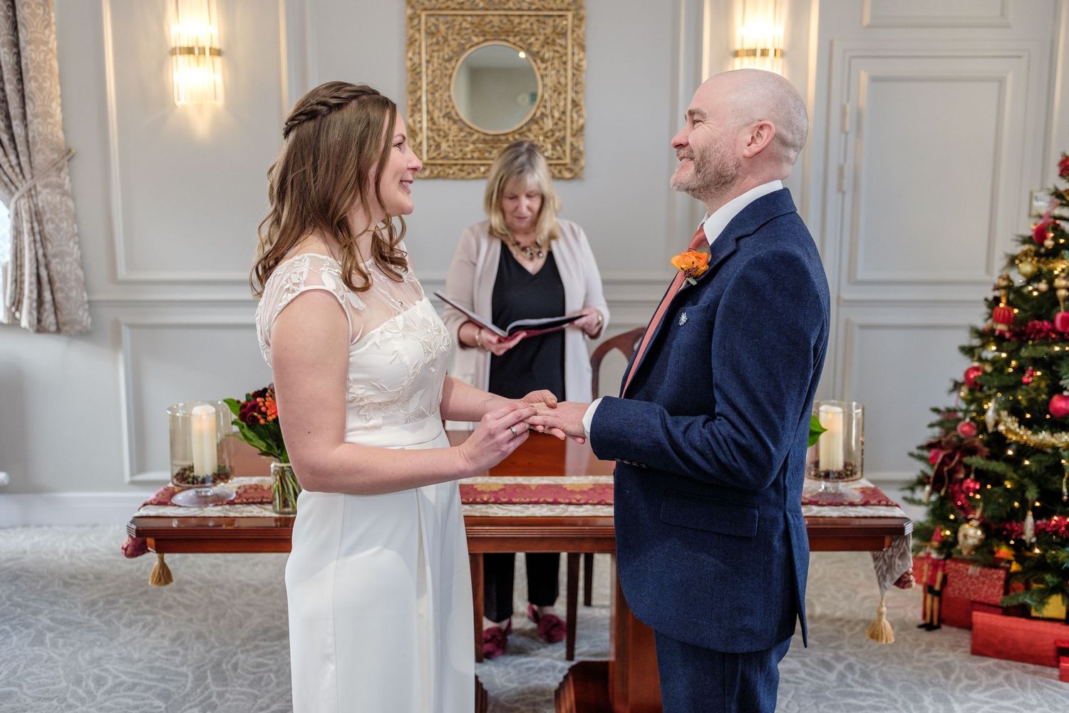 A couple exchanges rings during an elegant winter wedding ceremony in a bright white room decorated with Christmas trees.