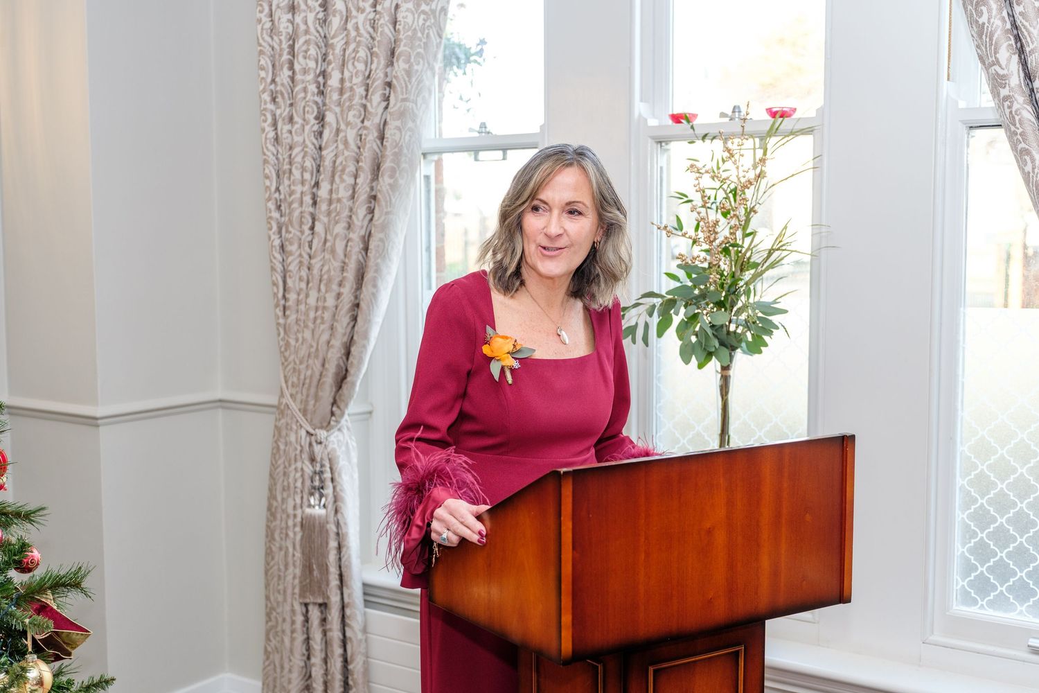 A person in a burgundy dress stands at a wooden podium in an elegant room with curtained windows and holiday decor.