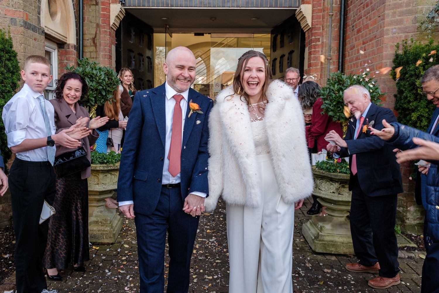 A group of wedding guests celebrate and applaud outside a brick church building during a joyful reception.