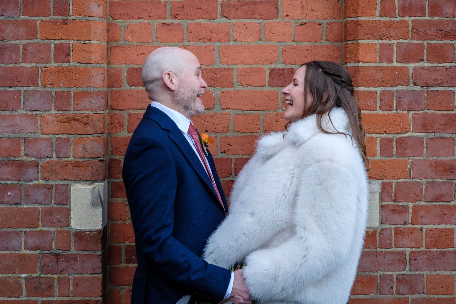 A couple shares a joyful moment together against a red brick wall, with the bride wearing a white fur coat.