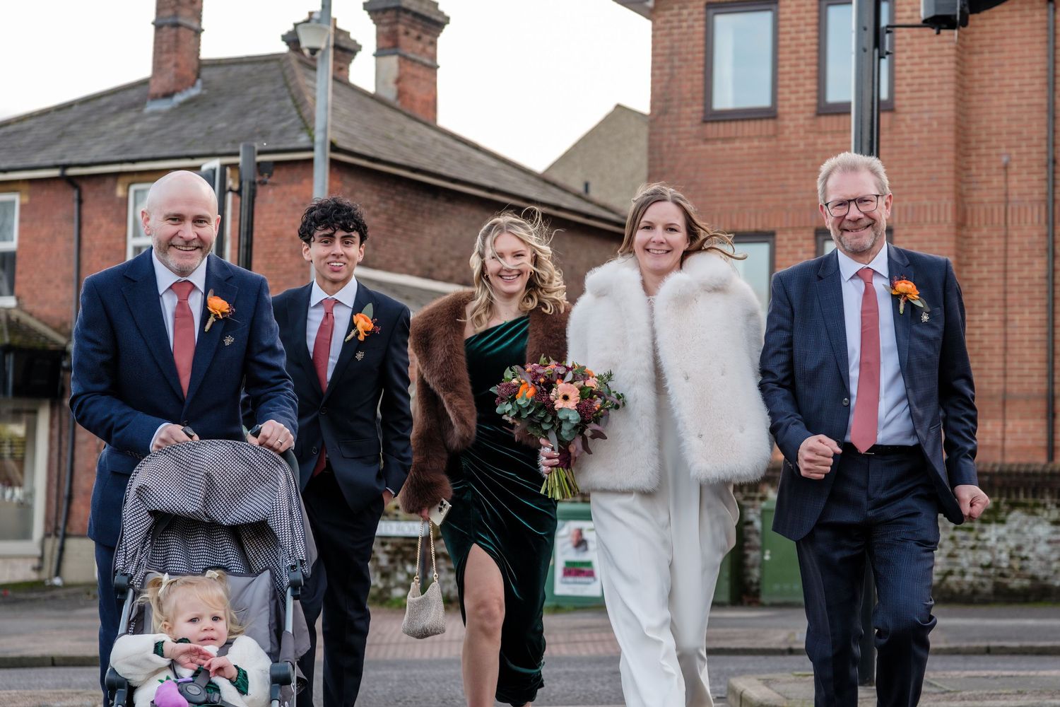 A wedding party walking together on a sidewalk with red brick buildings in the background on a cloudy day.