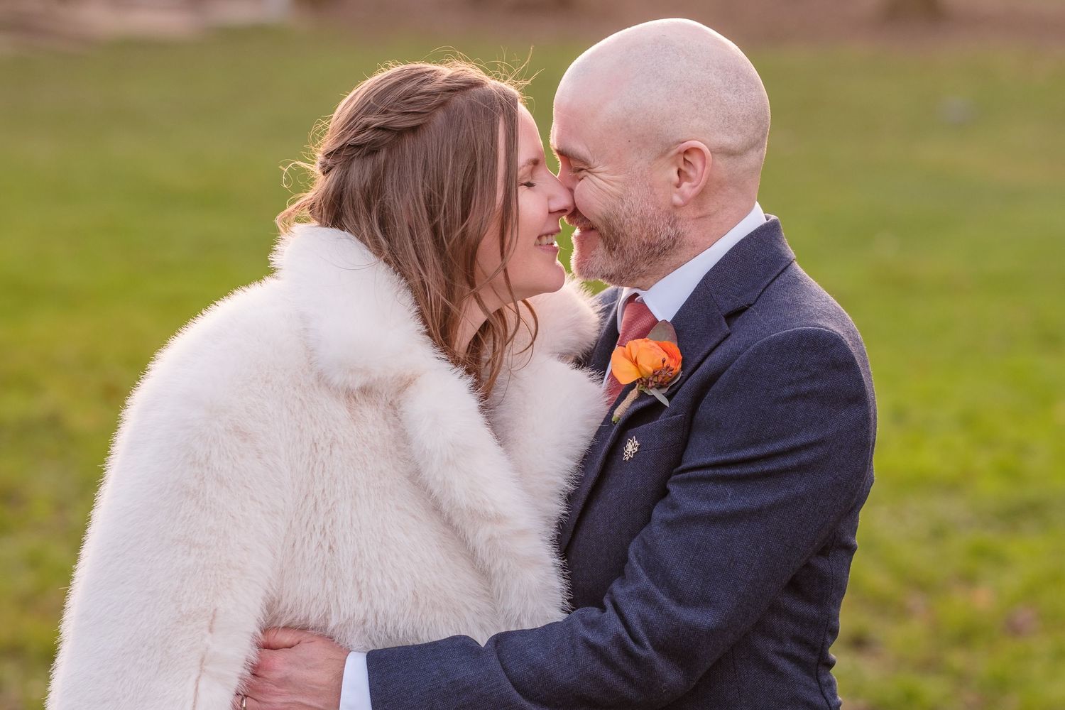 A romantic wedding couple shares tender moments in a grassy field while wearing winter attire with white fur shawl details.