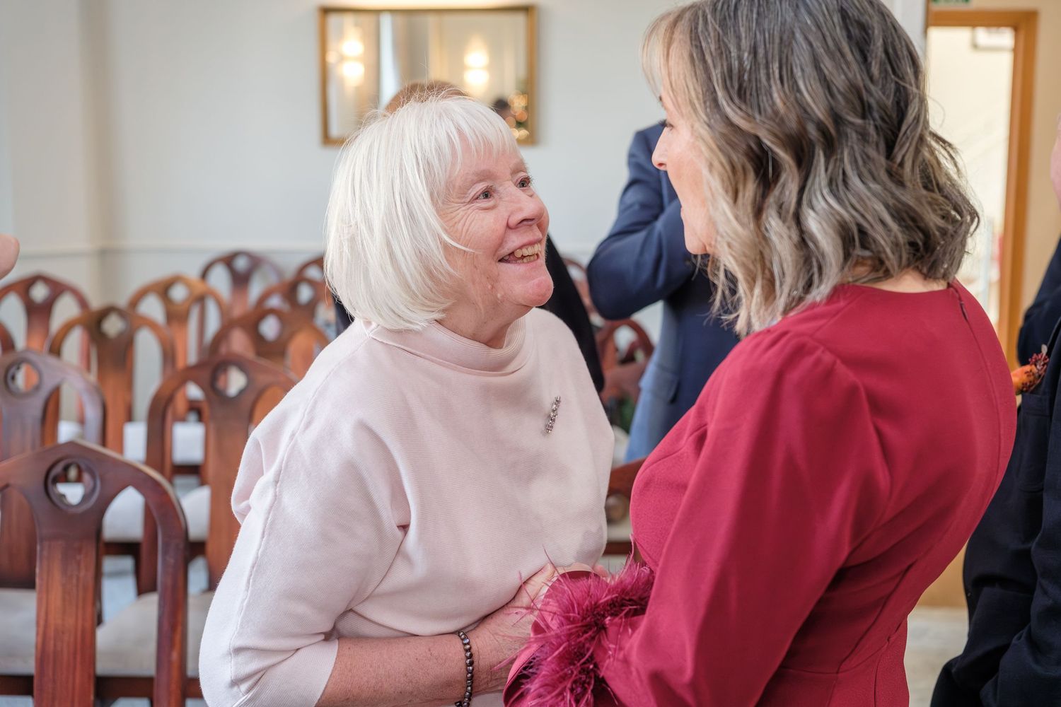 Two women in conversation at an indoor gathering, one wearing pink and one wearing red.