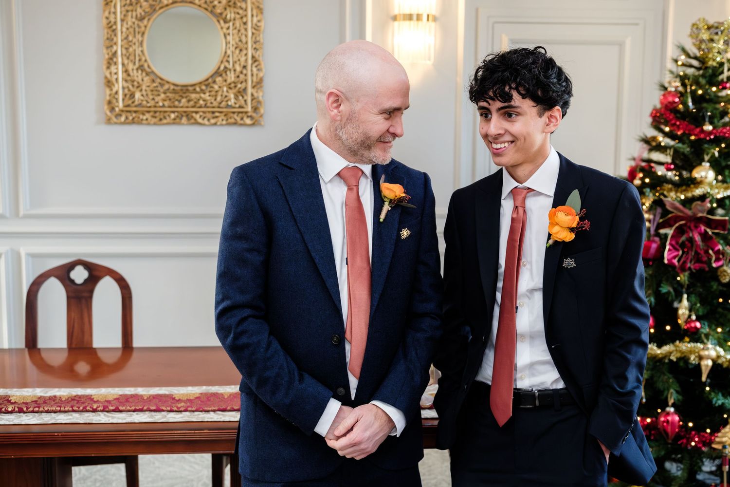 Two people in suits and red ties converse in an elegant room decorated for Christmas with a tree in the background.