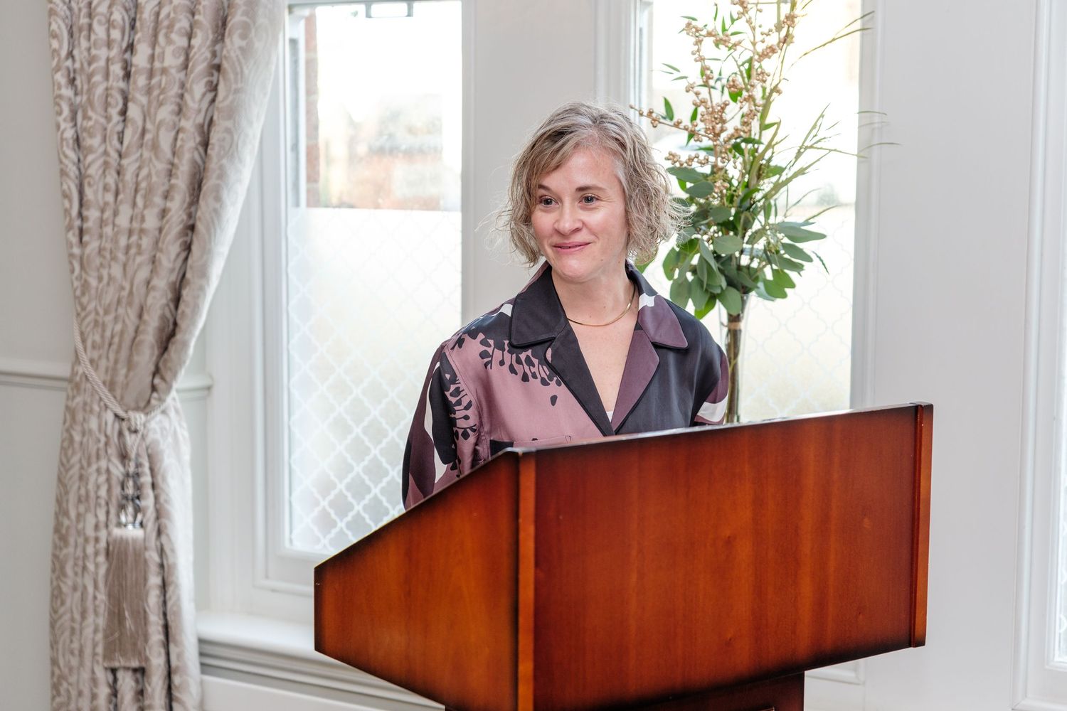 A woman in a pink shirt plays a wooden upright piano in a bright room with curtained windows.