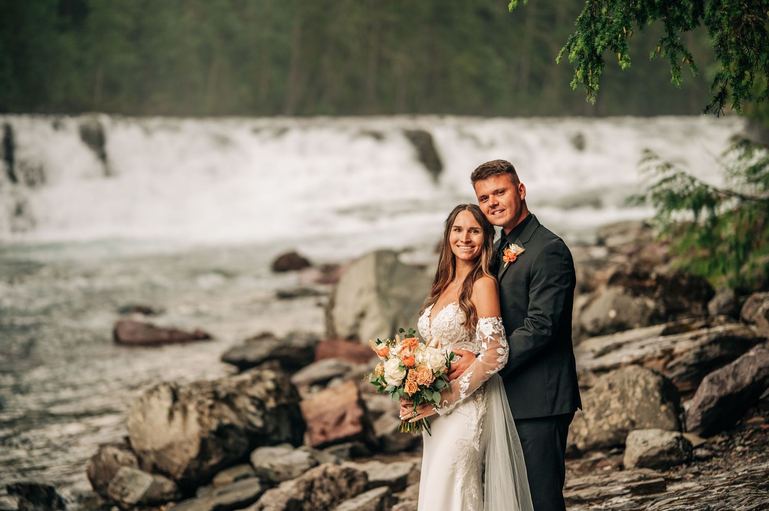 Couple in wedding attire embraces on rocky beach with rushing river in background during sunset photoshoot.