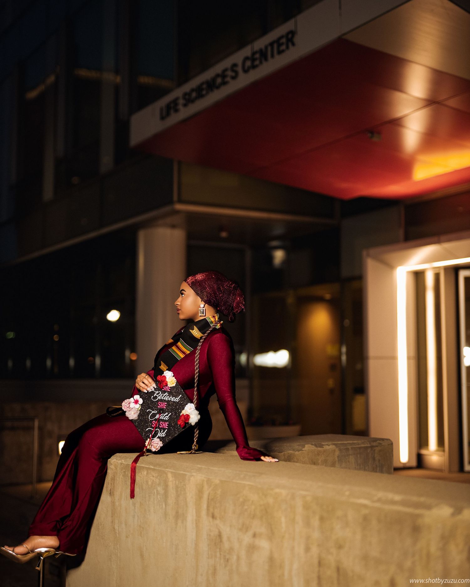 A person in a red coat lies dramatically posed across a concrete ledge at night with urban lighting in the background.