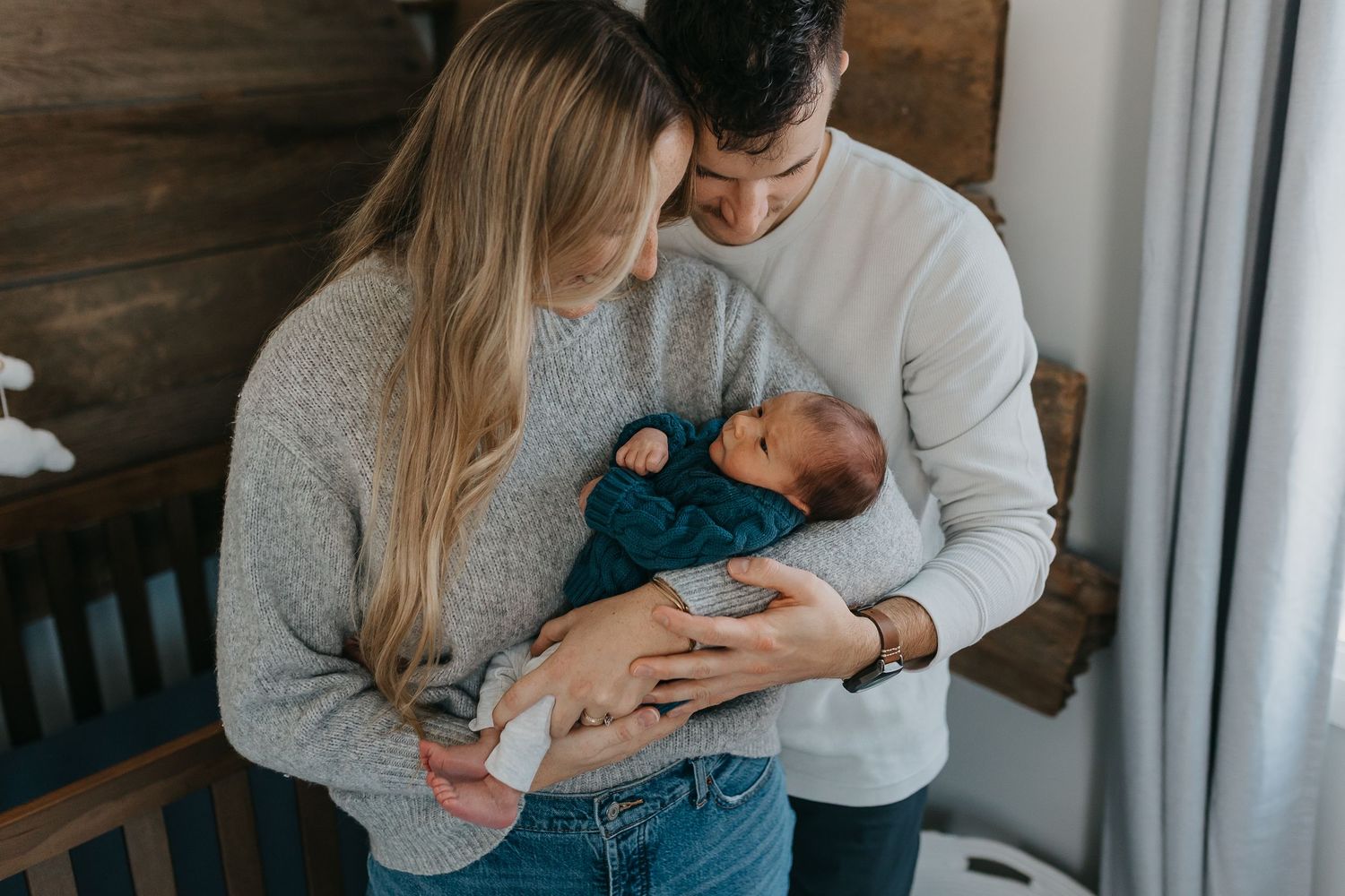 A couple tenderly holds their newborn baby wearing teal clothing while standing by a window in a rustic room.