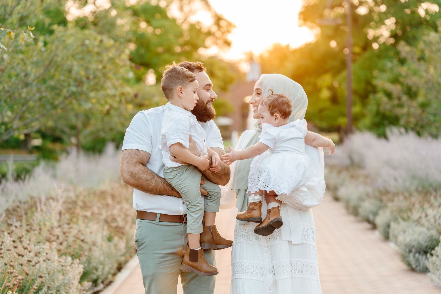 A happy Muslim family with two young children walks along a garden path in coordinated white and sage outfits at sunset.
