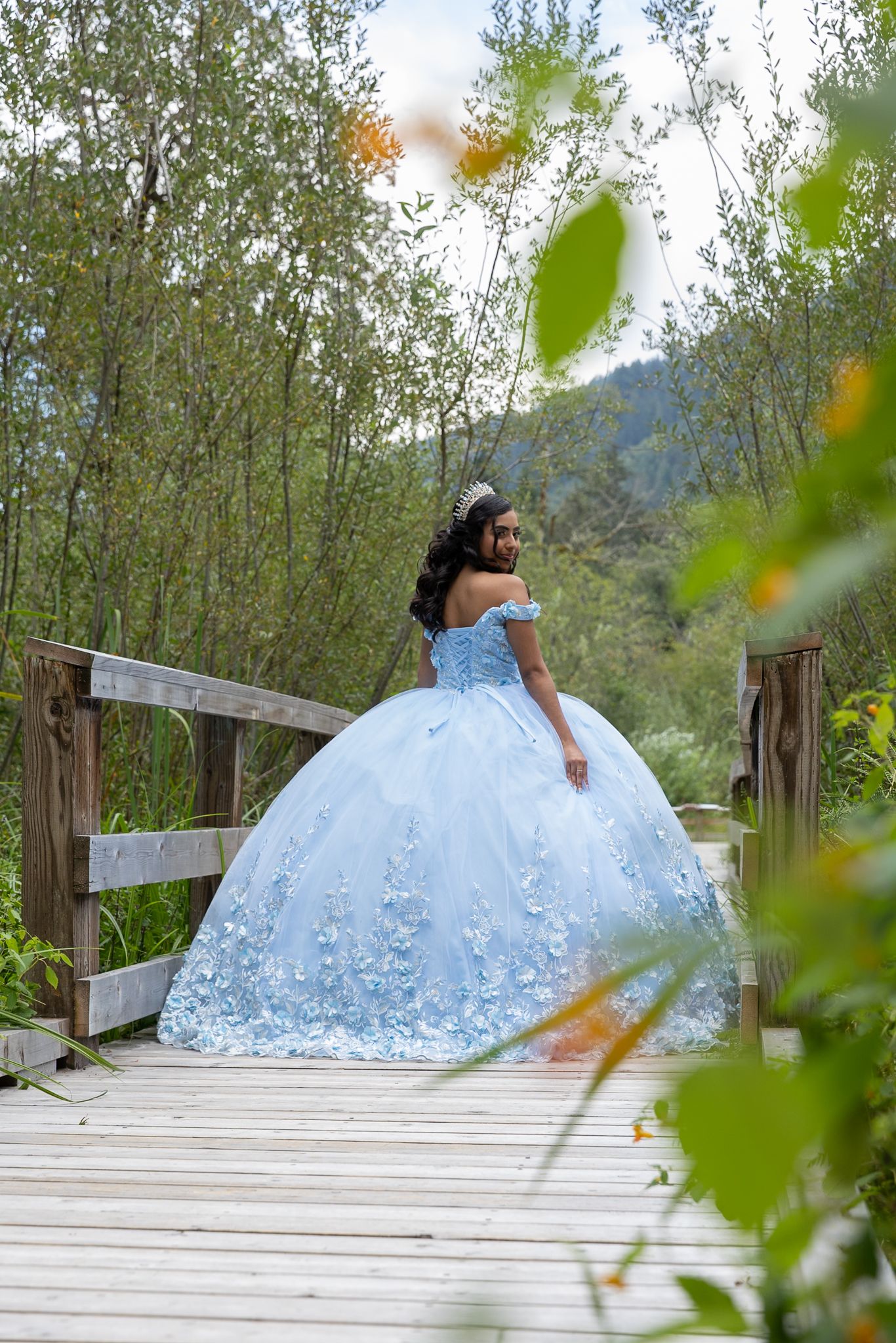 Elegant quinceañera in powder blue ball gown with 3D floral appliques poses on rustic wooden bridge in nature.