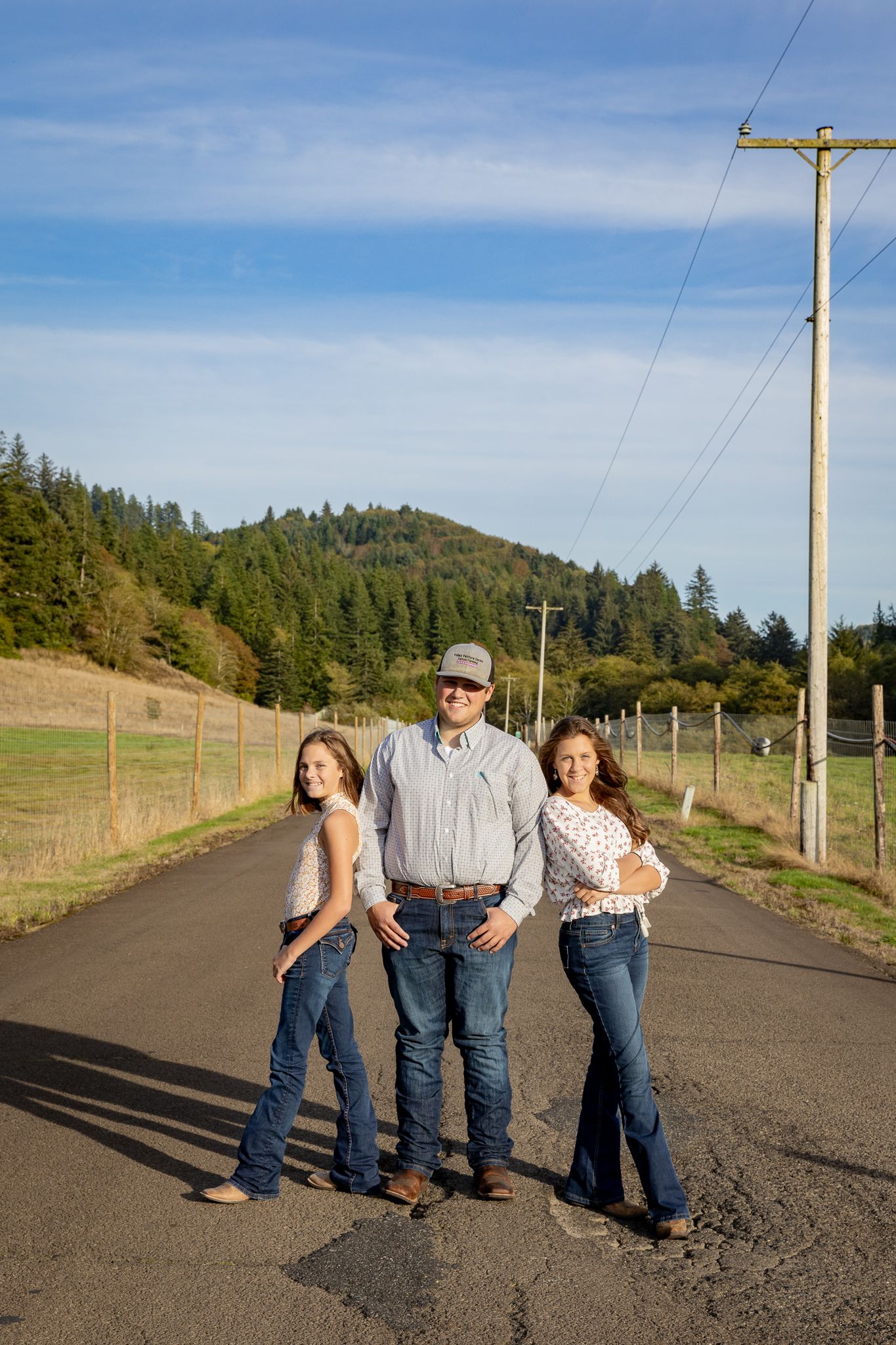 A family of three poses on a rural road surrounded by green hills and pine trees under a blue sky.