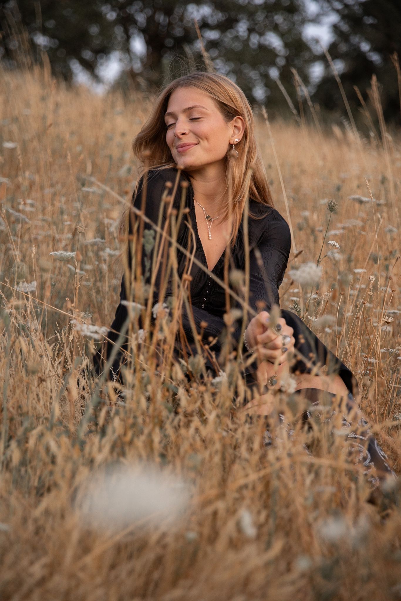 A blonde woman sits peacefully in tall dry grass with eyes closed, enjoying nature.