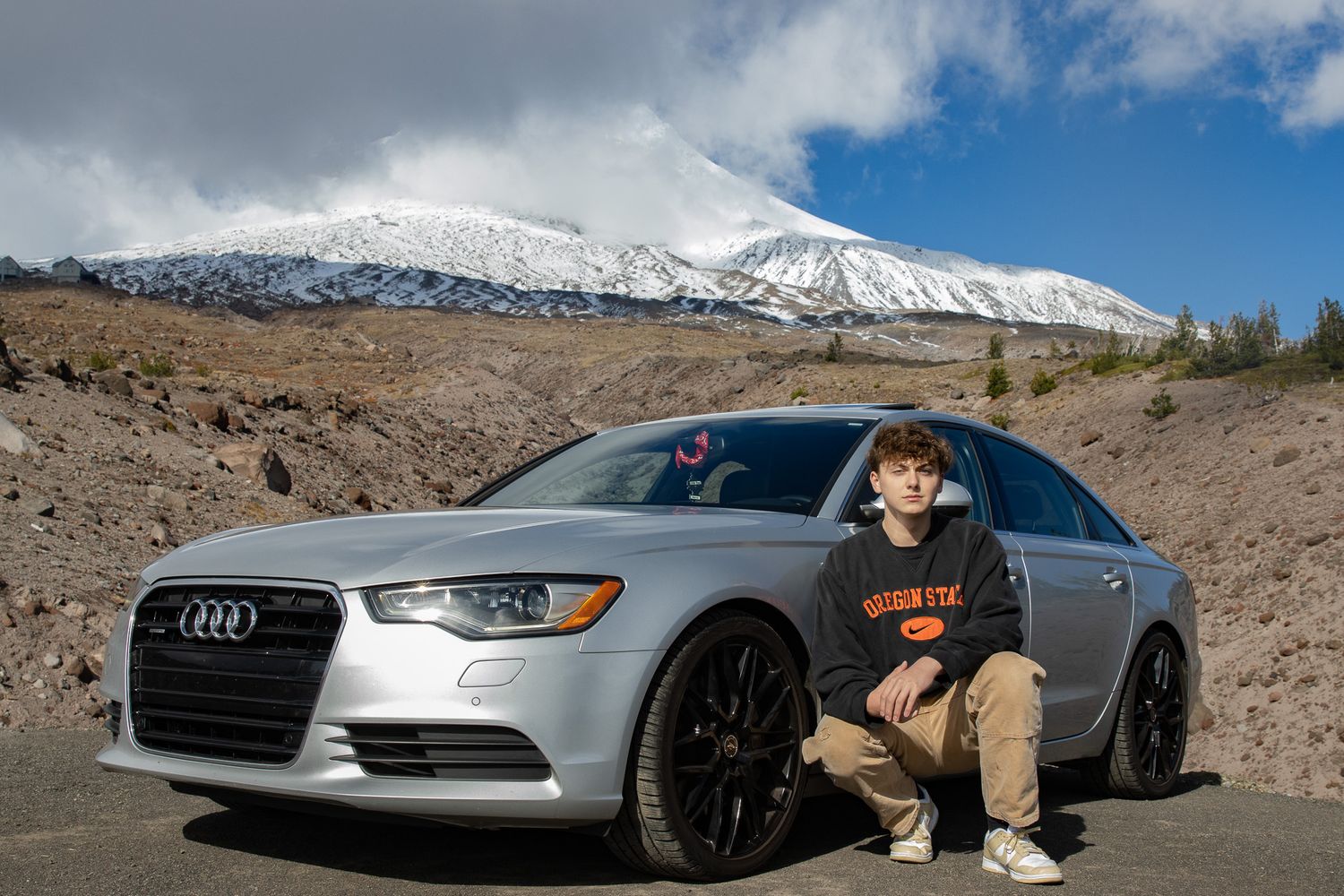 Young man crouching beside silver Audi A6 with snow-capped mount hood behind him.