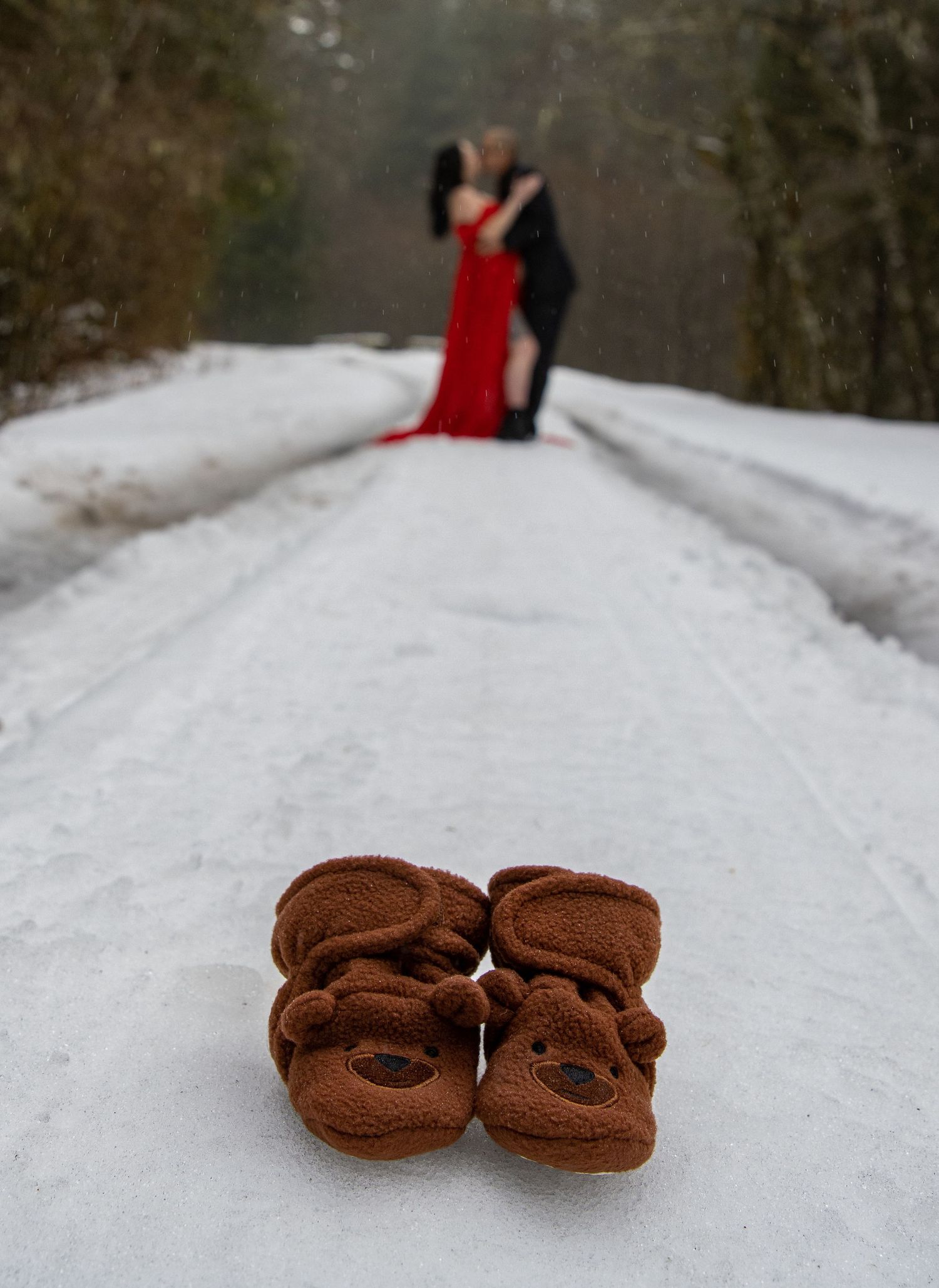 Couple embraces on a snowy road with adorable brown teddy bear baby booties in the foreground for maternity photos.