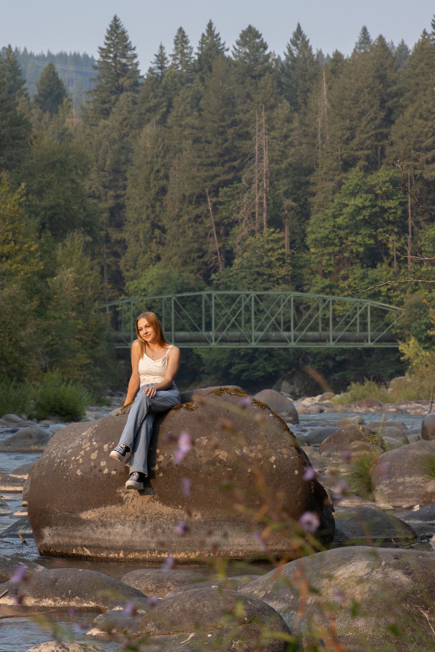 A teenage girl sits on a large boulder by a rocky river with a green bridge behind her.