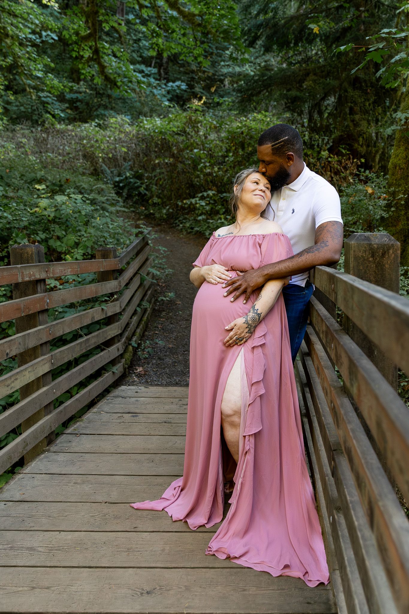 Couple embracing on wooden bridge, man holding pregnant woman's belly in lush green forest setting.