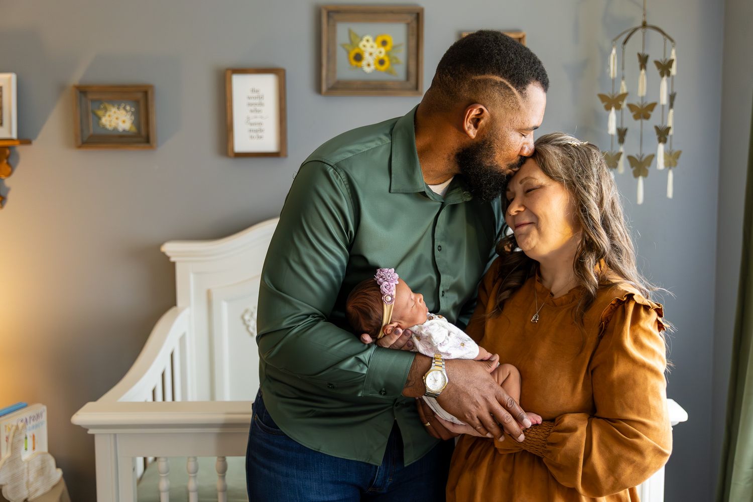 Interracial couple embracing while holding newborn baby near white crib.