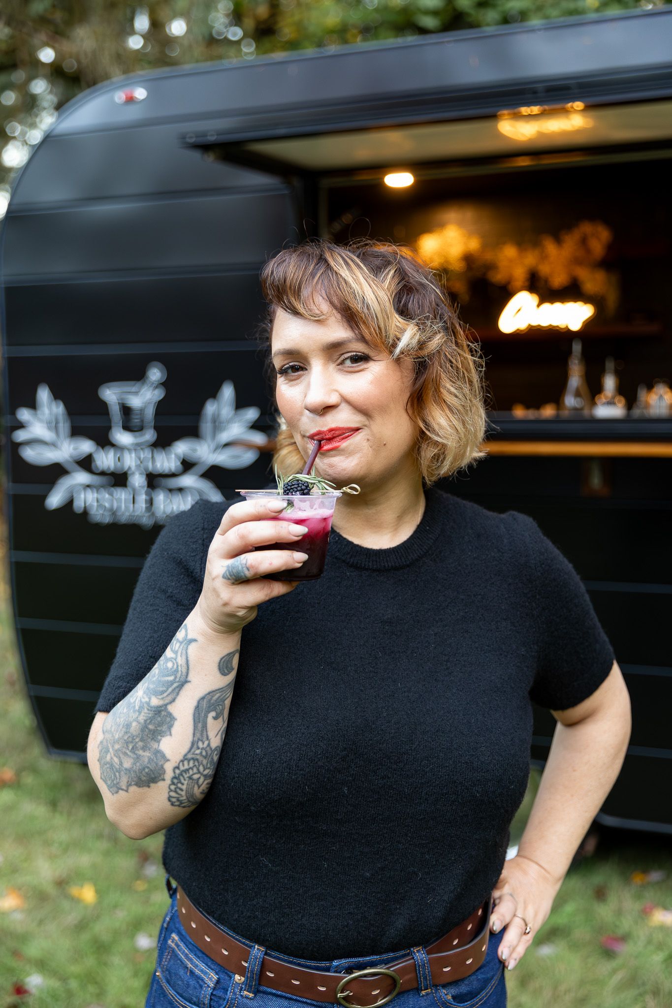Tattooed woman sipping a dark berry cocktail outside a black vintage mobile bar trailer.
