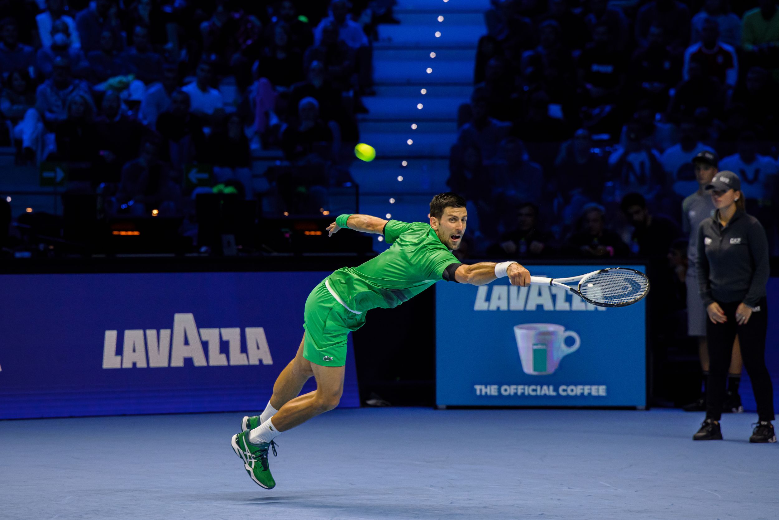 Tennis player in green outfit making a dynamic serve motion on blue indoor court.