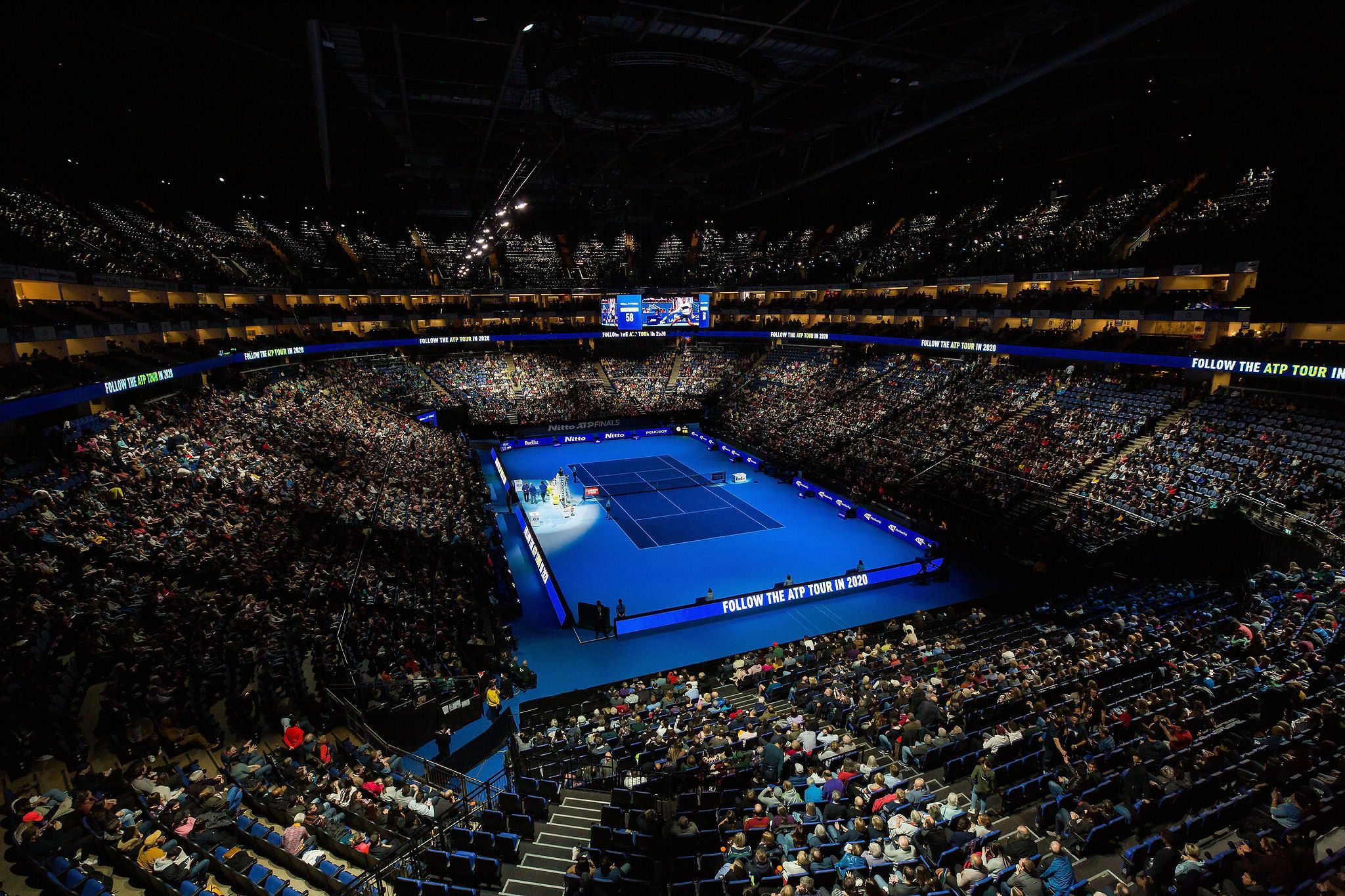 Tennis match at a packed indoor stadium with bright blue courts and stadium lighting.