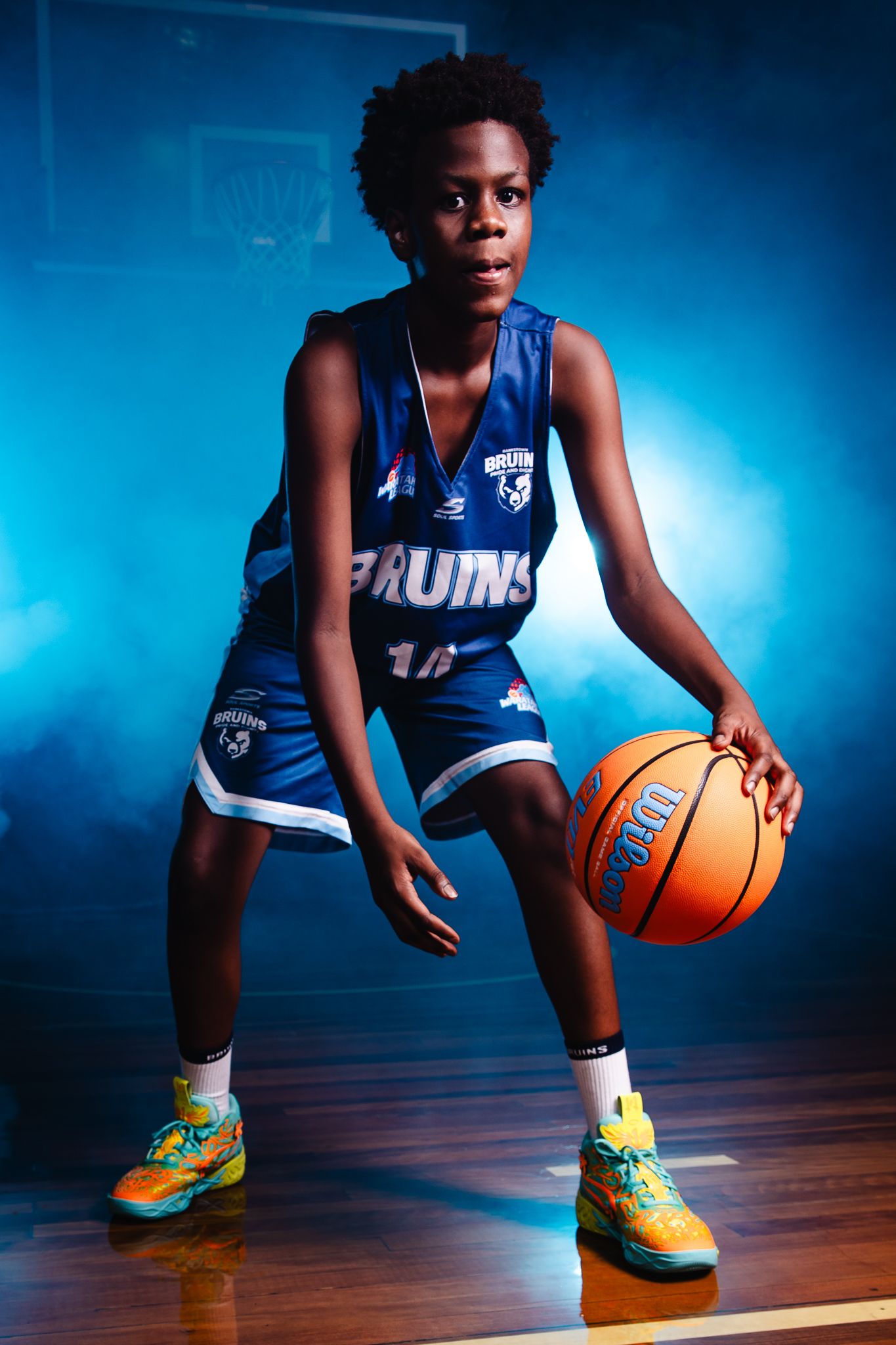 Basketball player in blue Bruins jersey dribbles on wooden court with dramatic blue lighting and colorful sneakers.