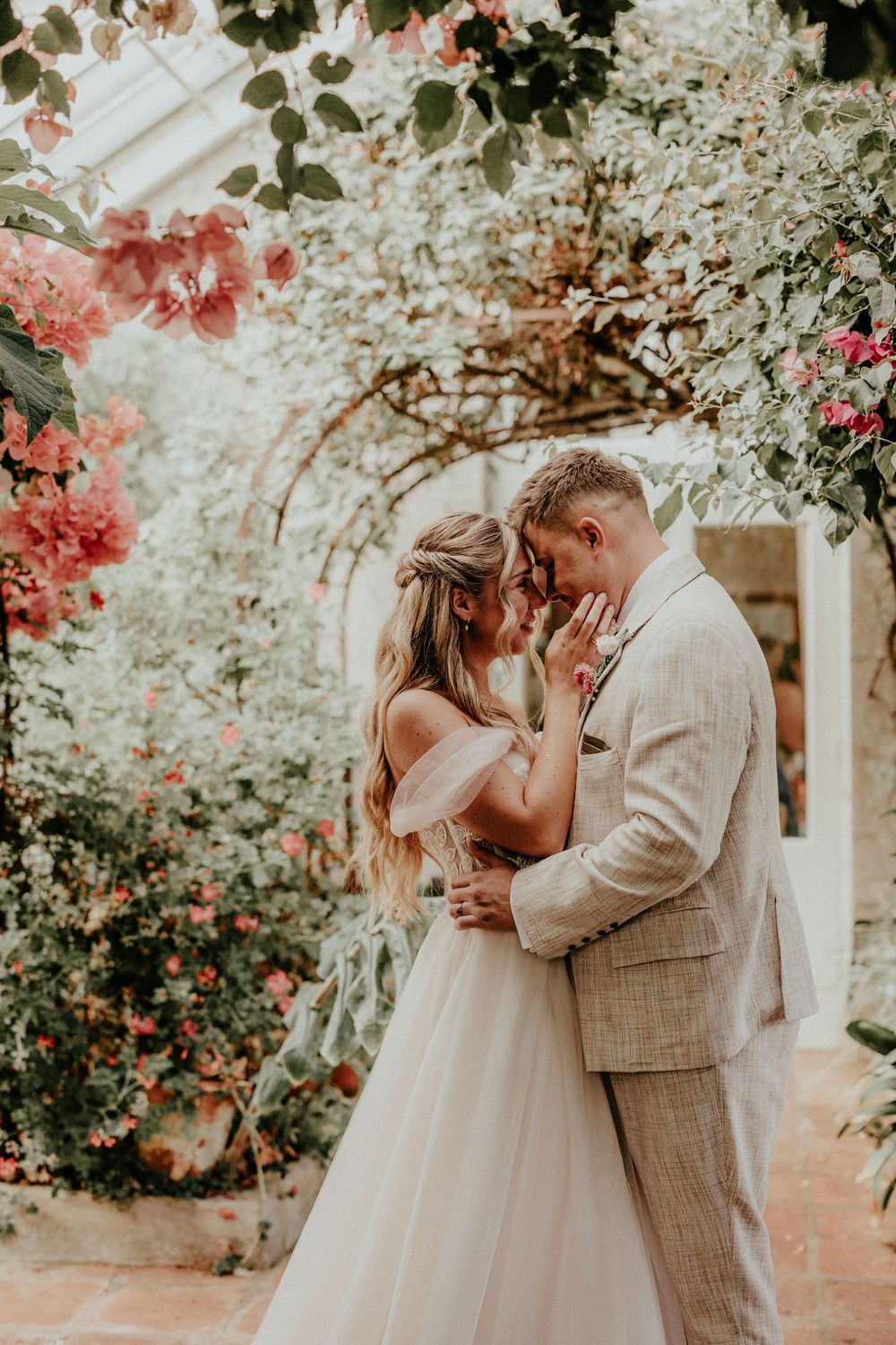 Romantic couple shares tender kiss under blooming bougainvillea flowers in garden wedding setting.