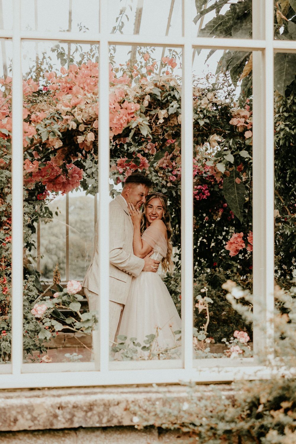 A romantic greenhouse wedding photo with pink bougainvillea flowers framing a couple through vintage window panes.
