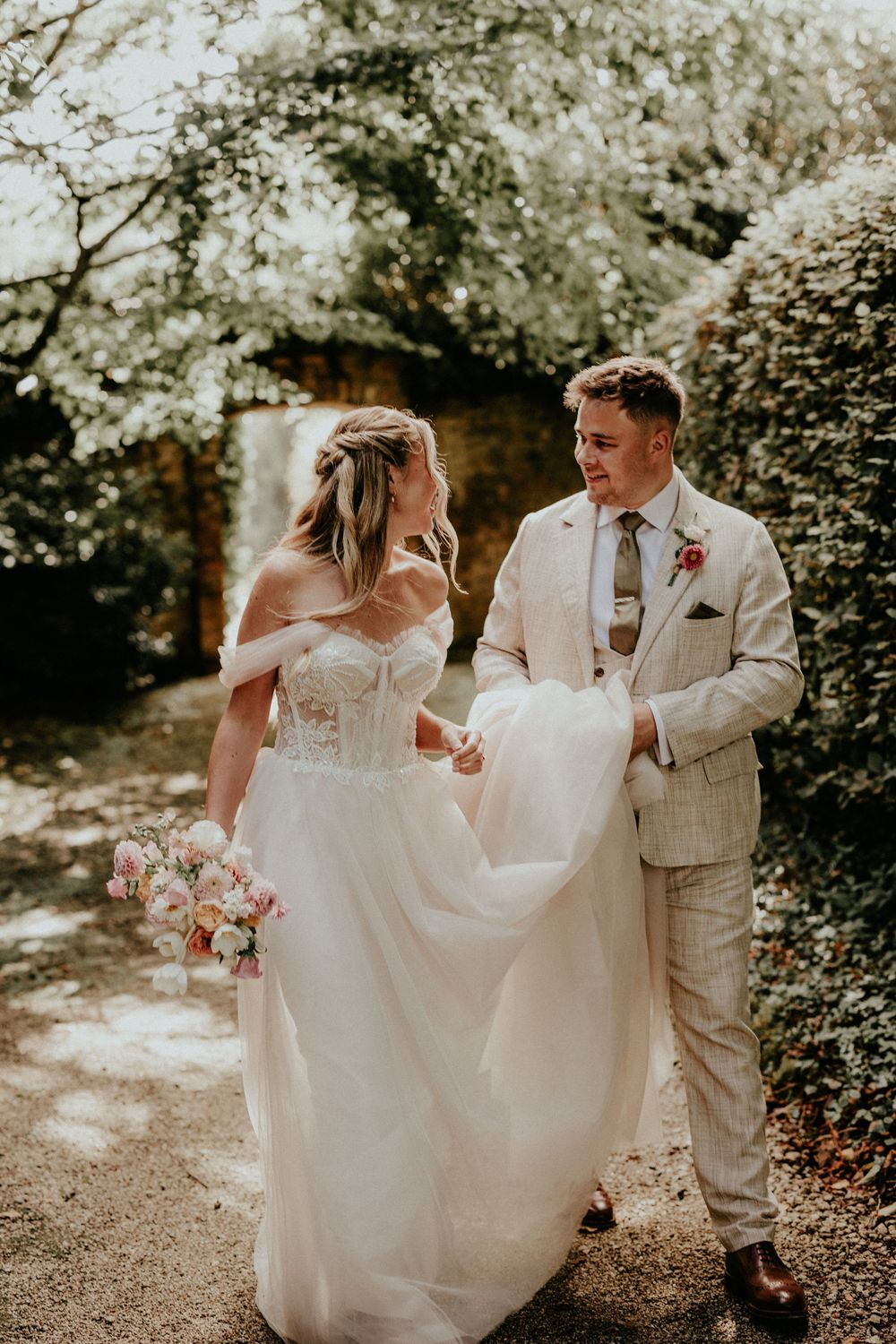 A couple shares a joyful moment on their wedding day in a sunlit garden path, wearing light coloured formal attire. Cornwall wedding photo