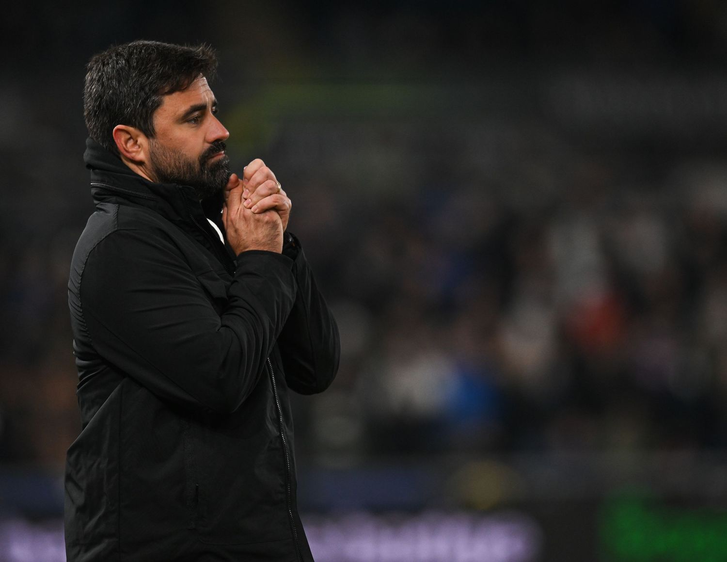 Soccer manager in black coat gestures from sideline during intense match action.