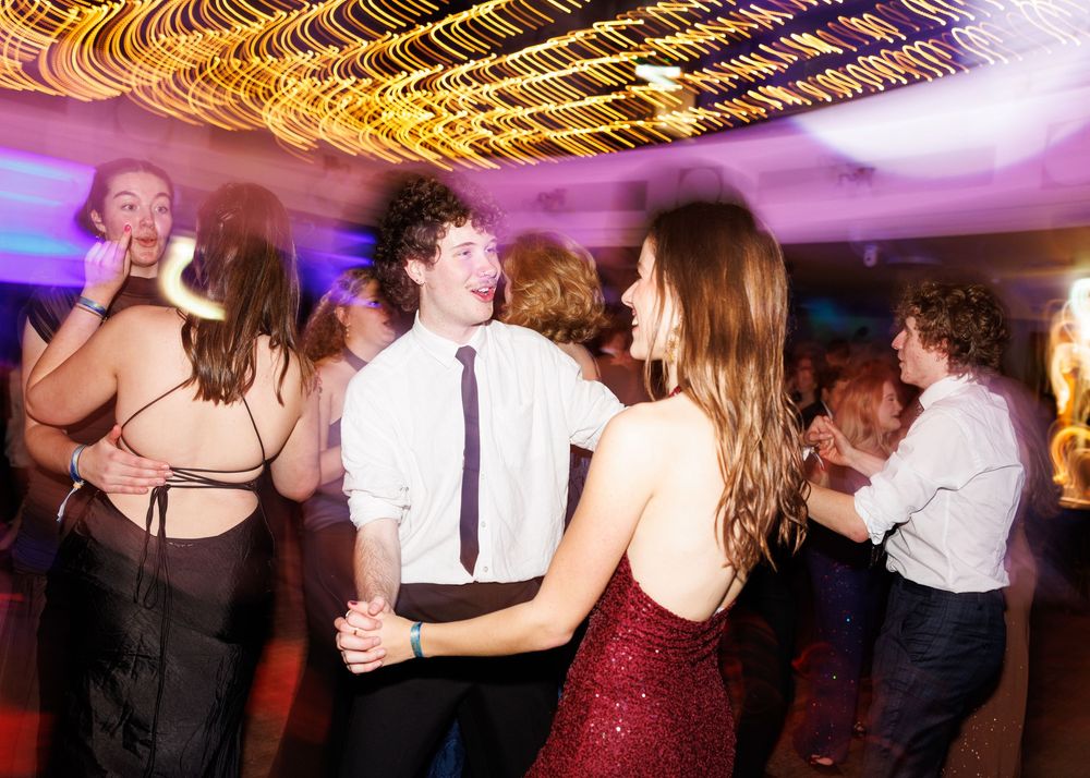 Couples dance together on a dimly lit dance floor with purple and golden lighting at an evening celebration.