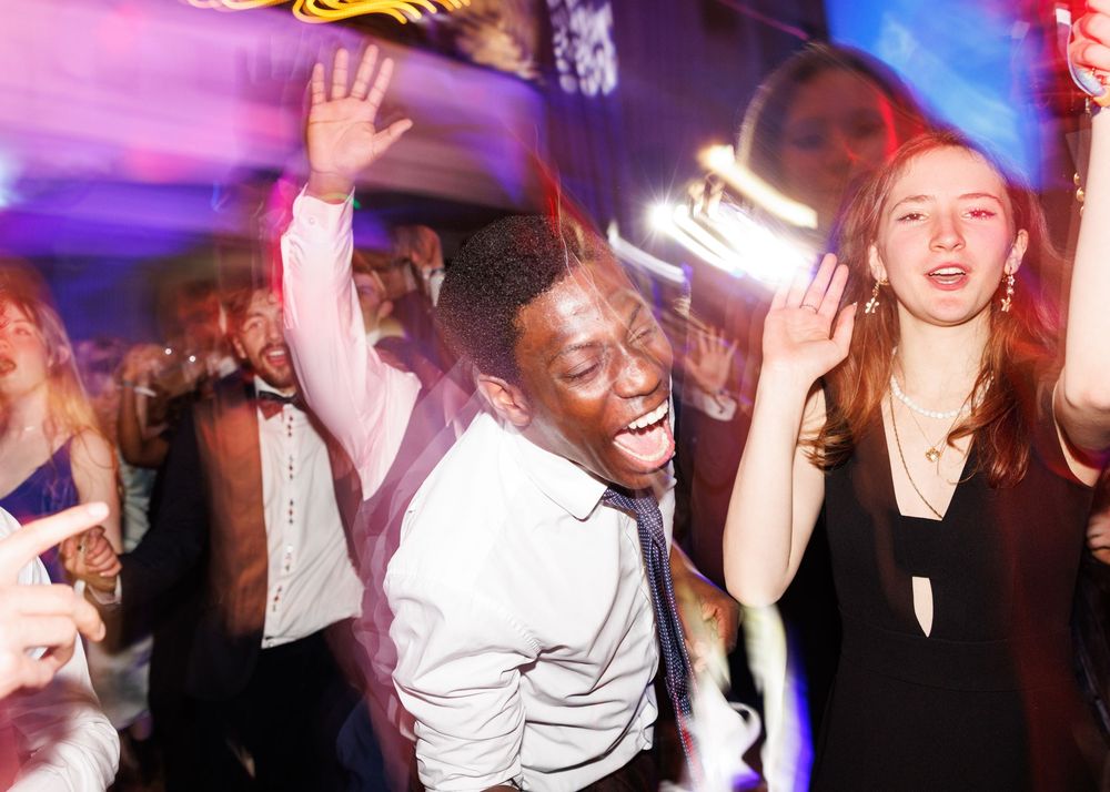 Crowd of people dancing and celebrating at a party with colorful purple and red lighting effects.