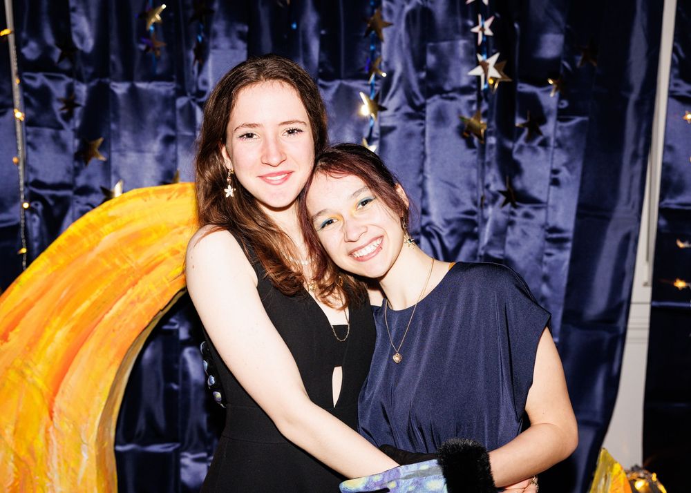 Two people pose together in front of a dark background with bright orange decor visible on the right side.