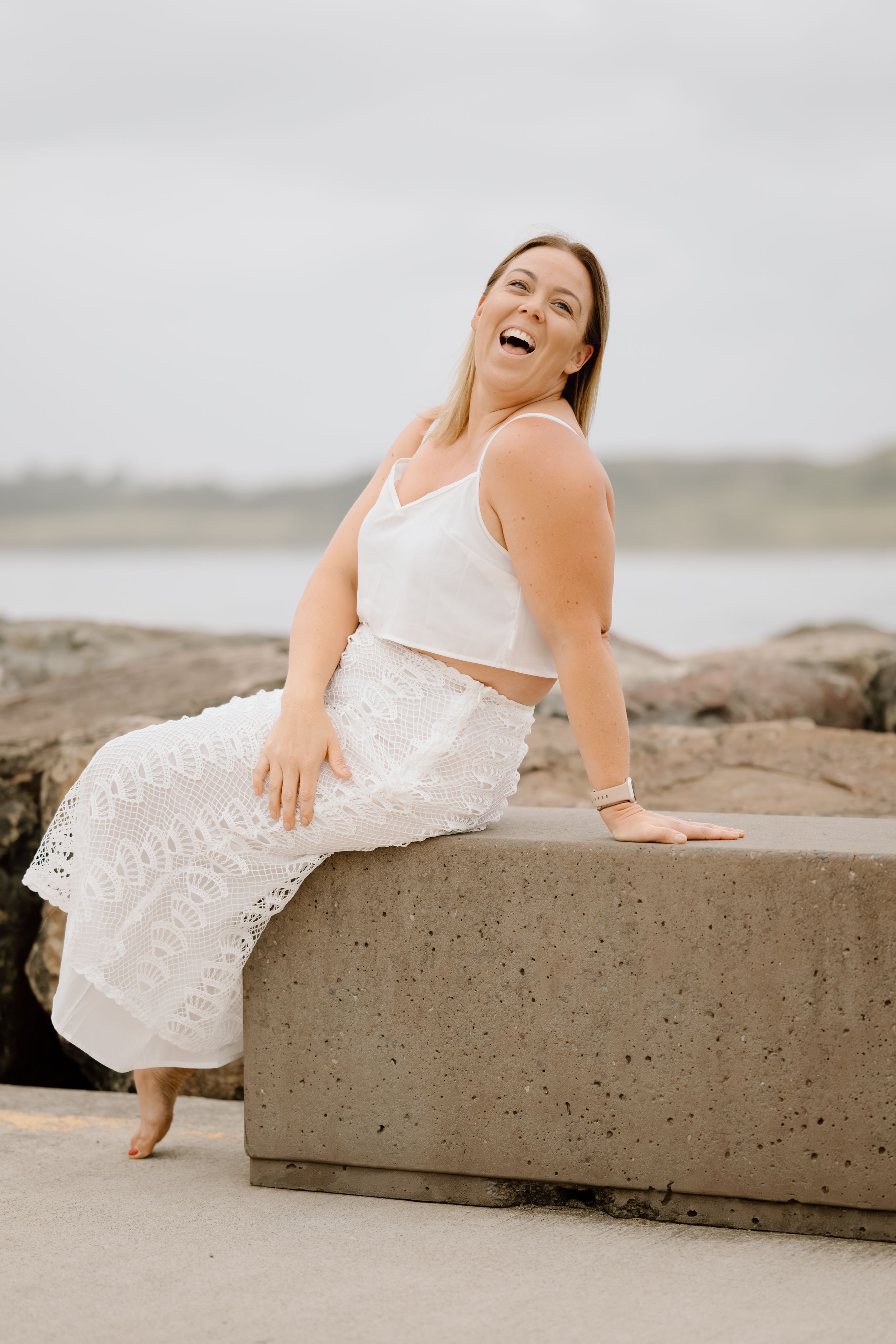 Joyful woman in white crop top and lace pants sits on concrete barrier at beach, laughing with genuine happiness.