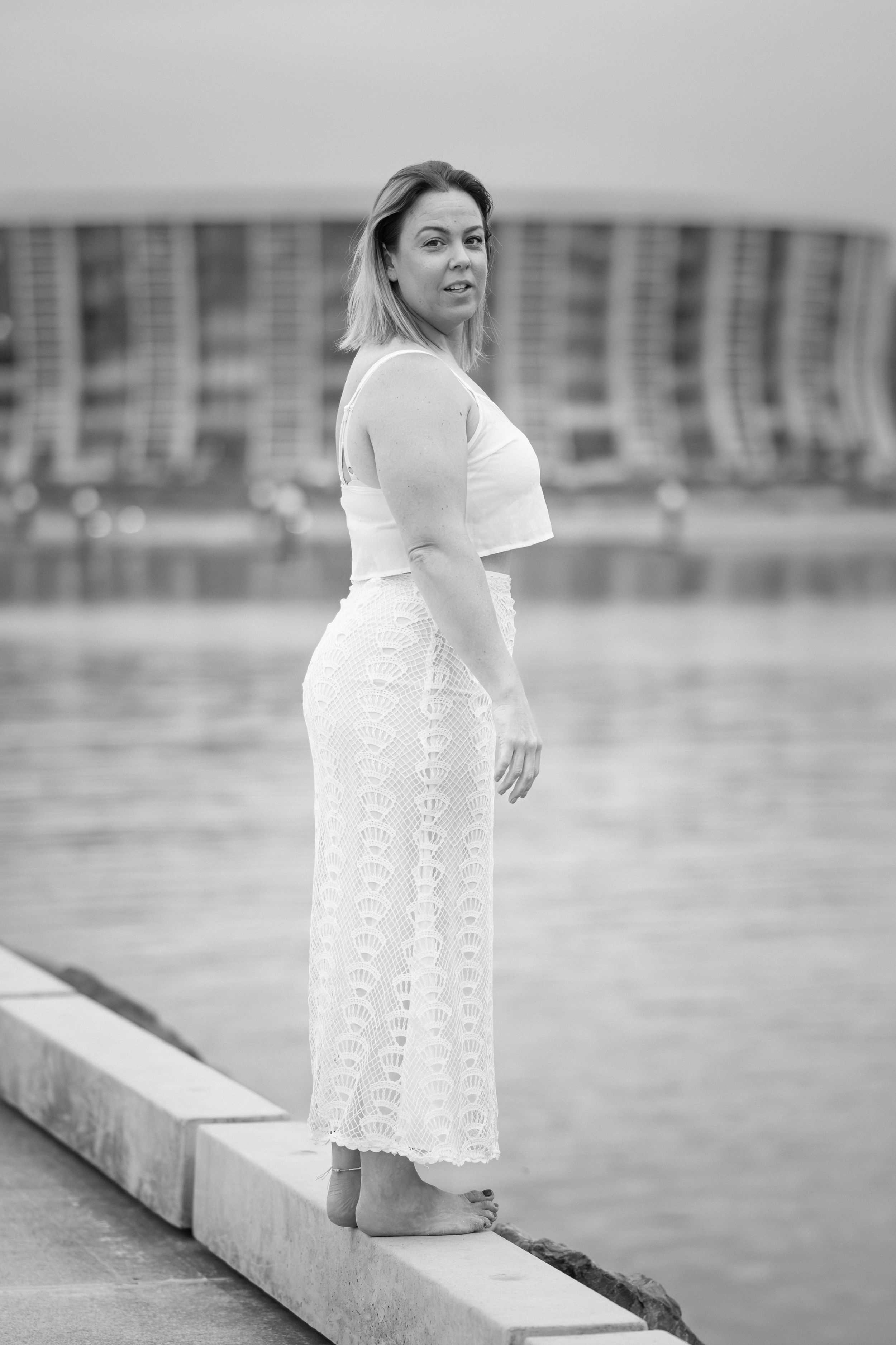 A person in an elegant white outfit poses on a concrete ledge near water with buildings in the background.