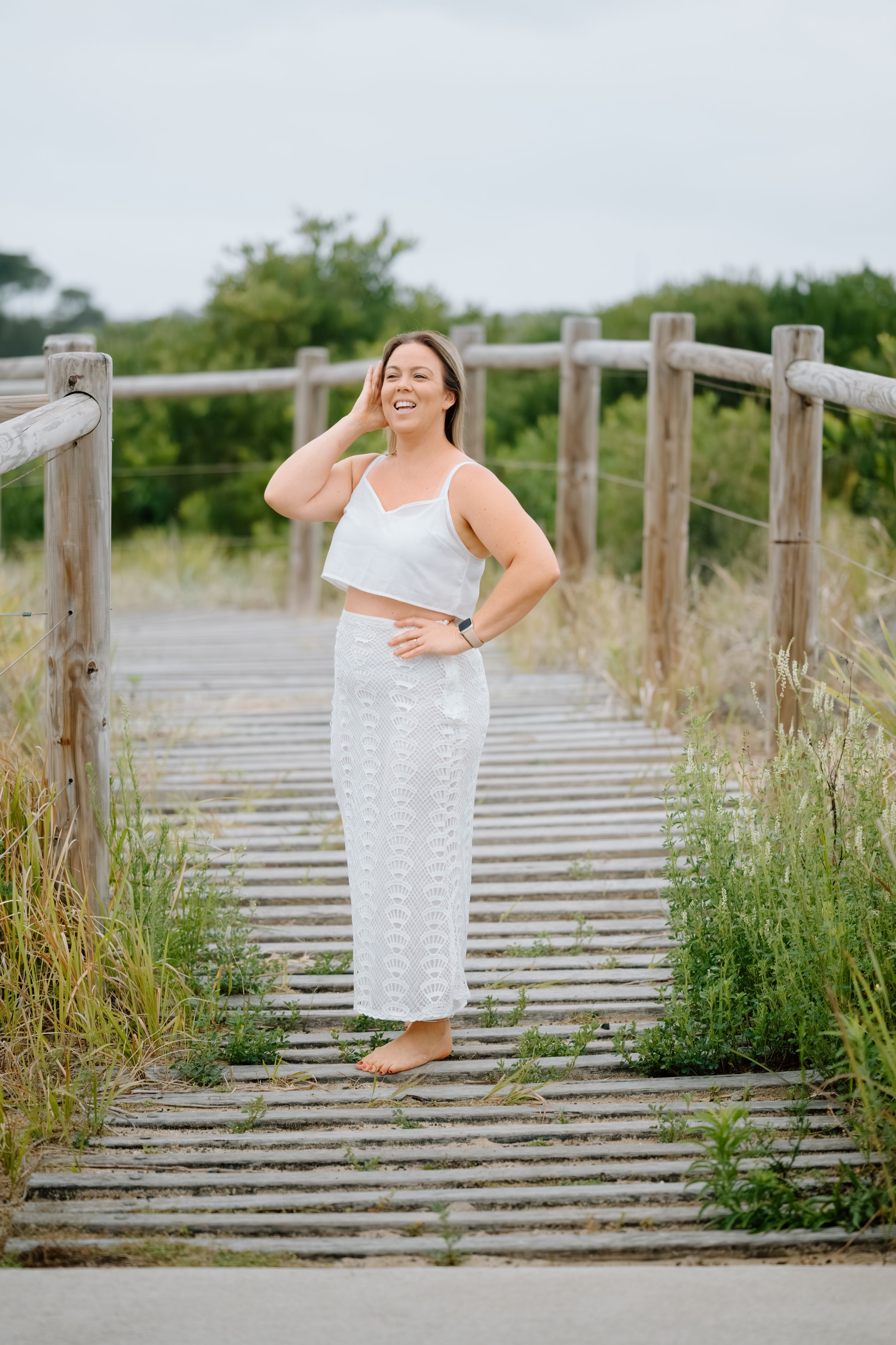 Person in white dress poses confidently on wooden boardwalk surrounded by coastal grass and natural scenery.
