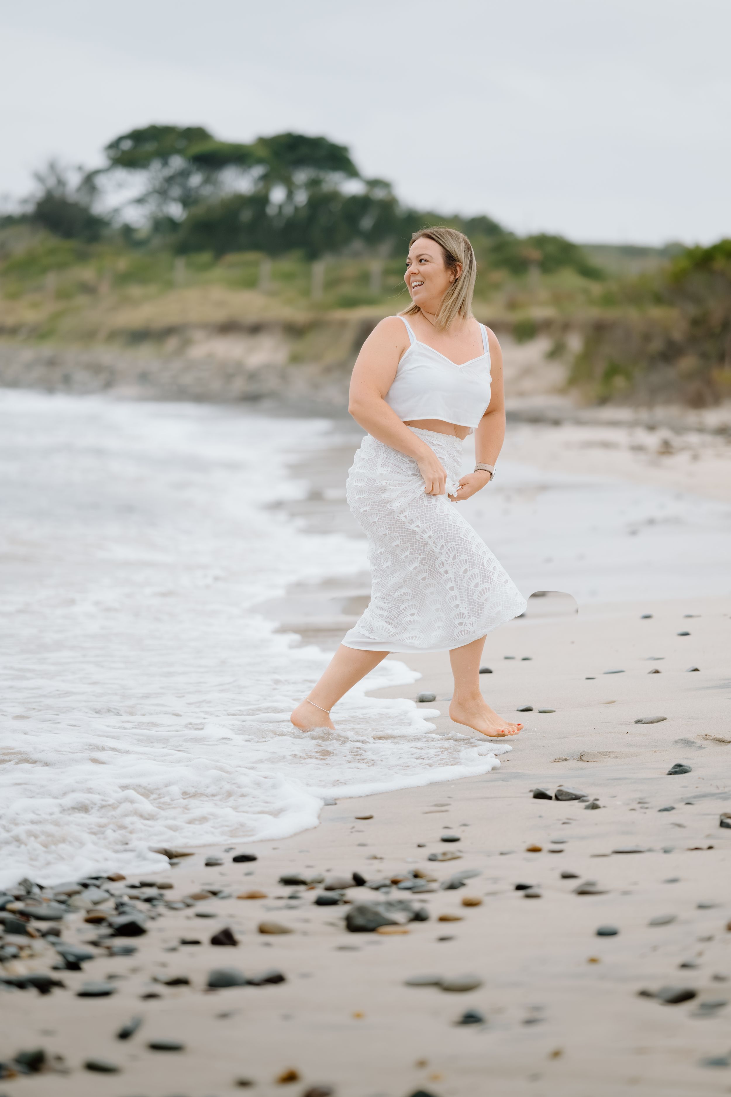 A person in a flowing white dress dances and twirls joyfully along a rocky beach shoreline.