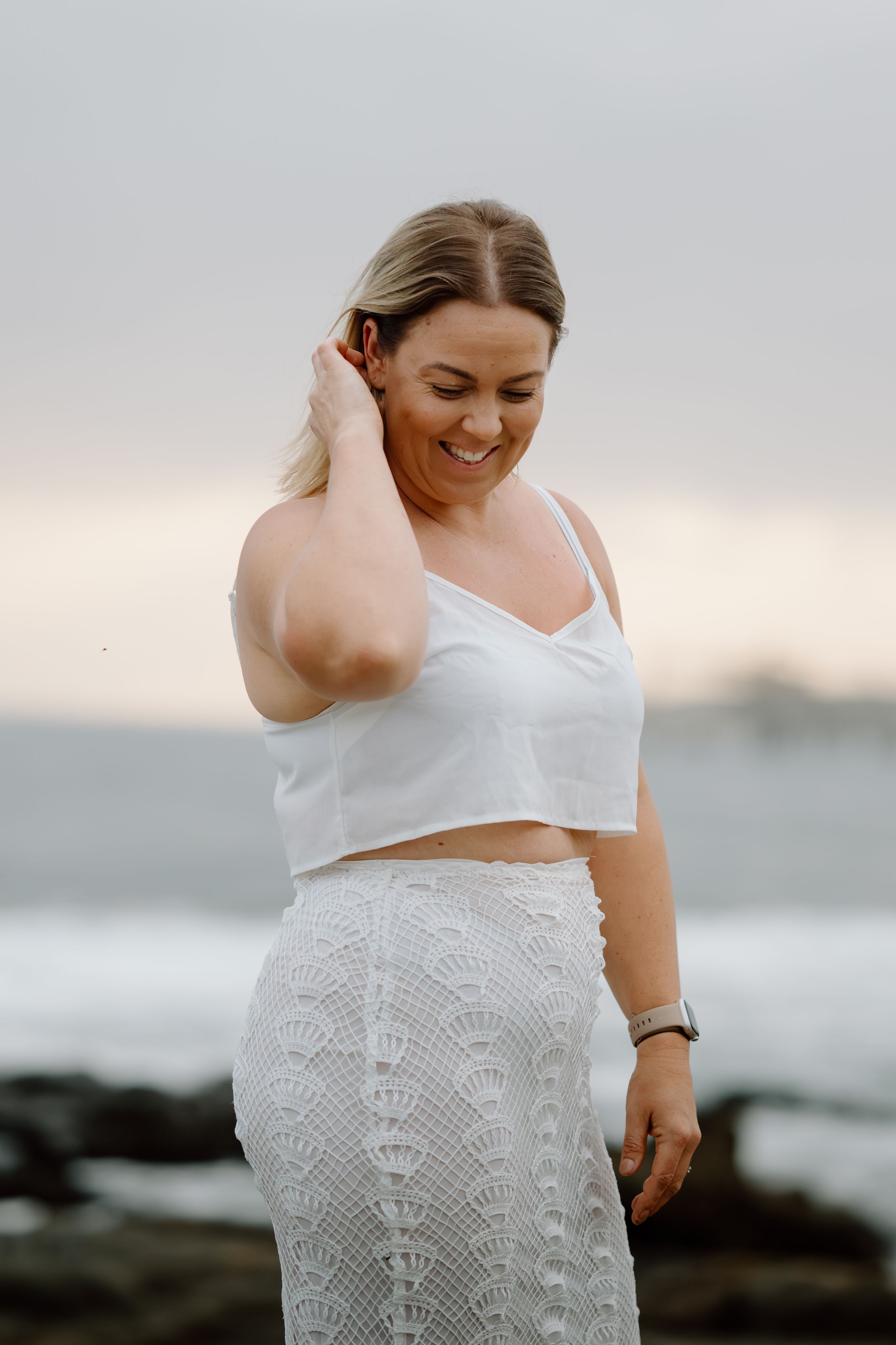 A person in a white lace dress poses by the ocean during golden hour at a rocky beach.
