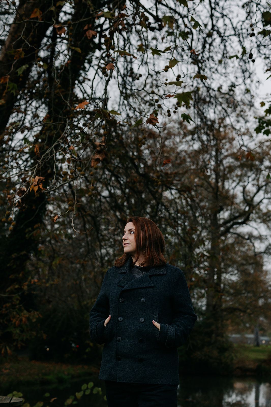 A person in a black sweater stands among bare autumn trees near a lake on an overcast day. Fantasy writer headshots