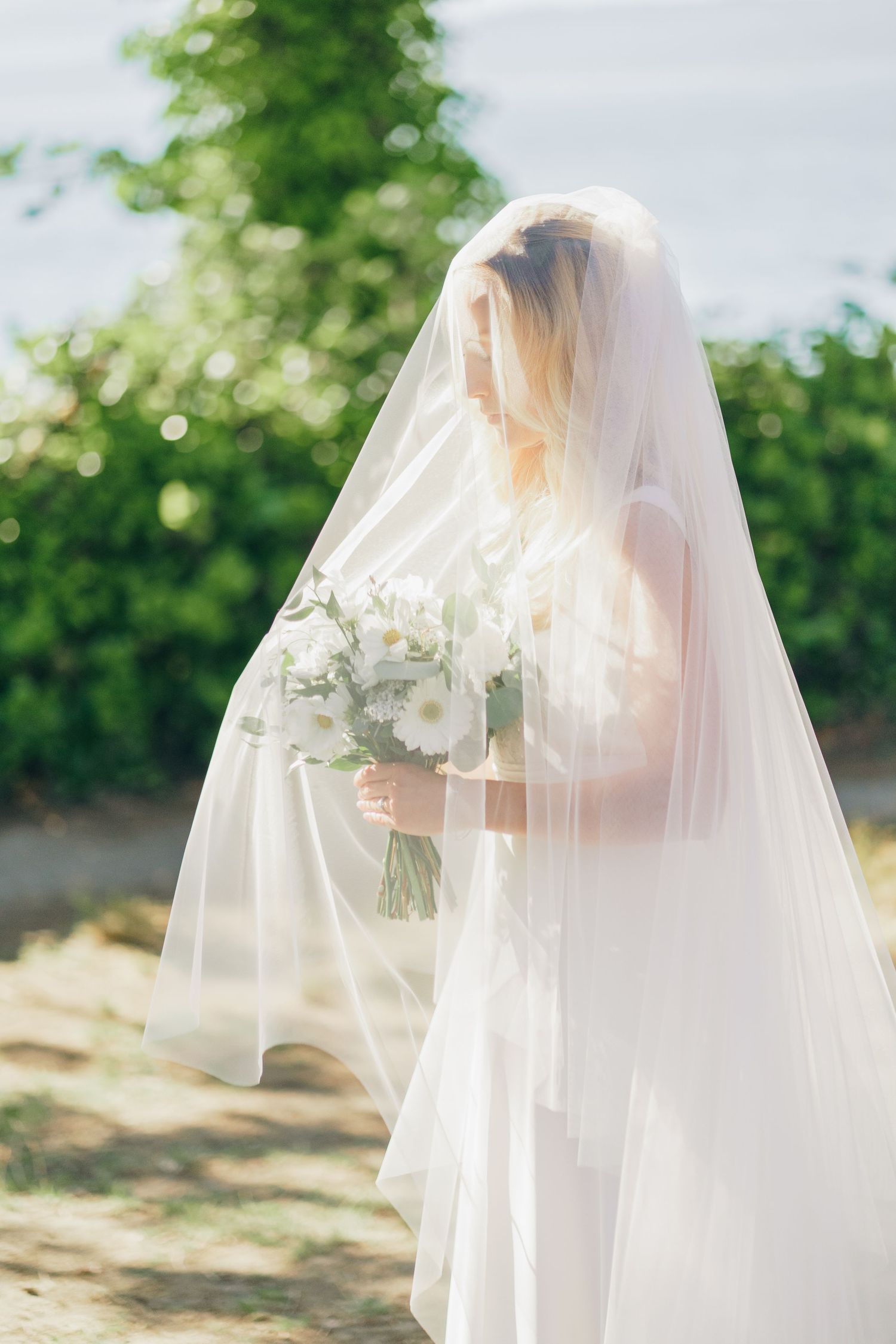 A bride in a flowing white veil holds a delicate bouquet of white flowers against a sunlit garden backdrop.