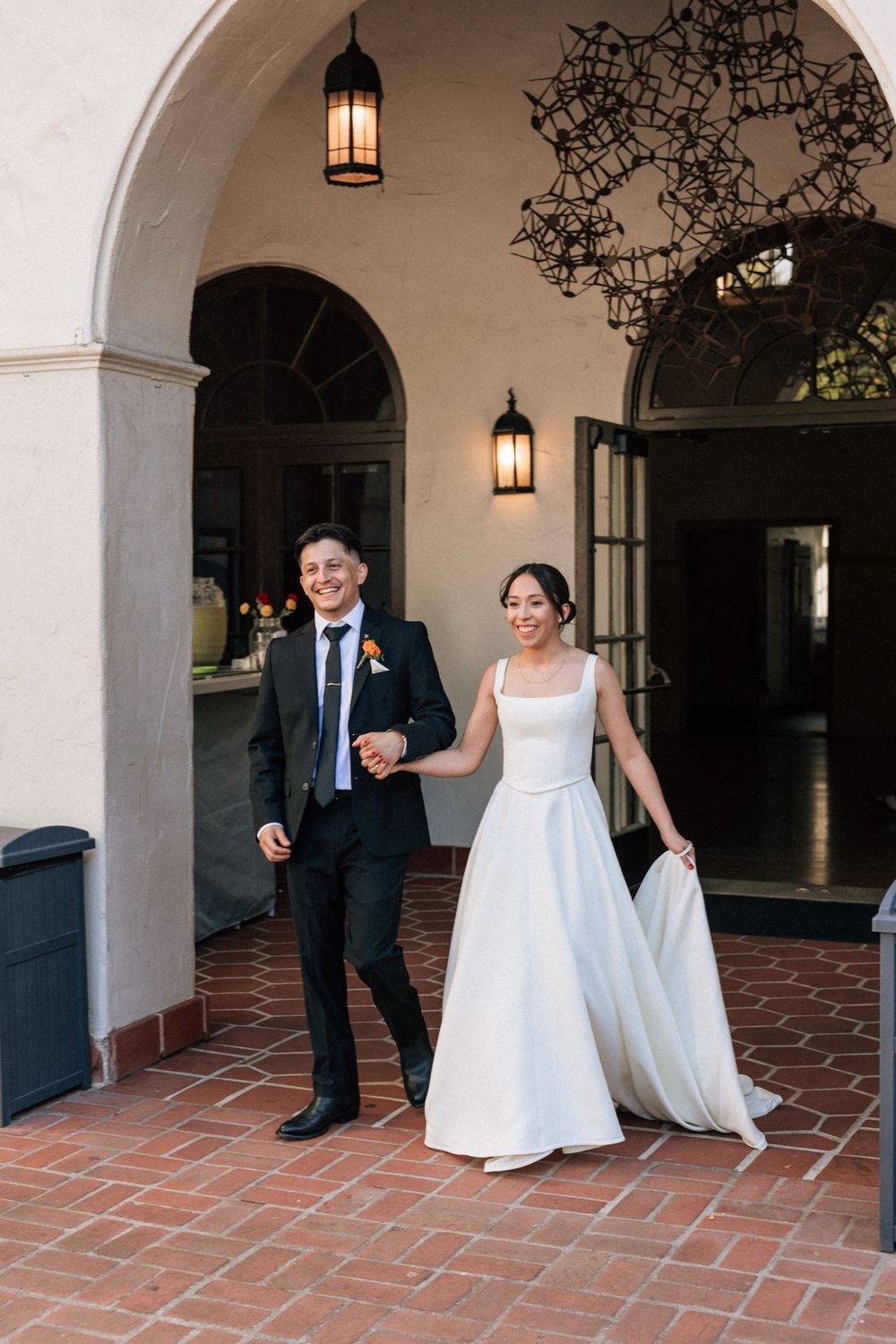 A bride and groom hold hands and smile walking in to greet their guests at their Unitarian Society of Santa Barbara wedding.