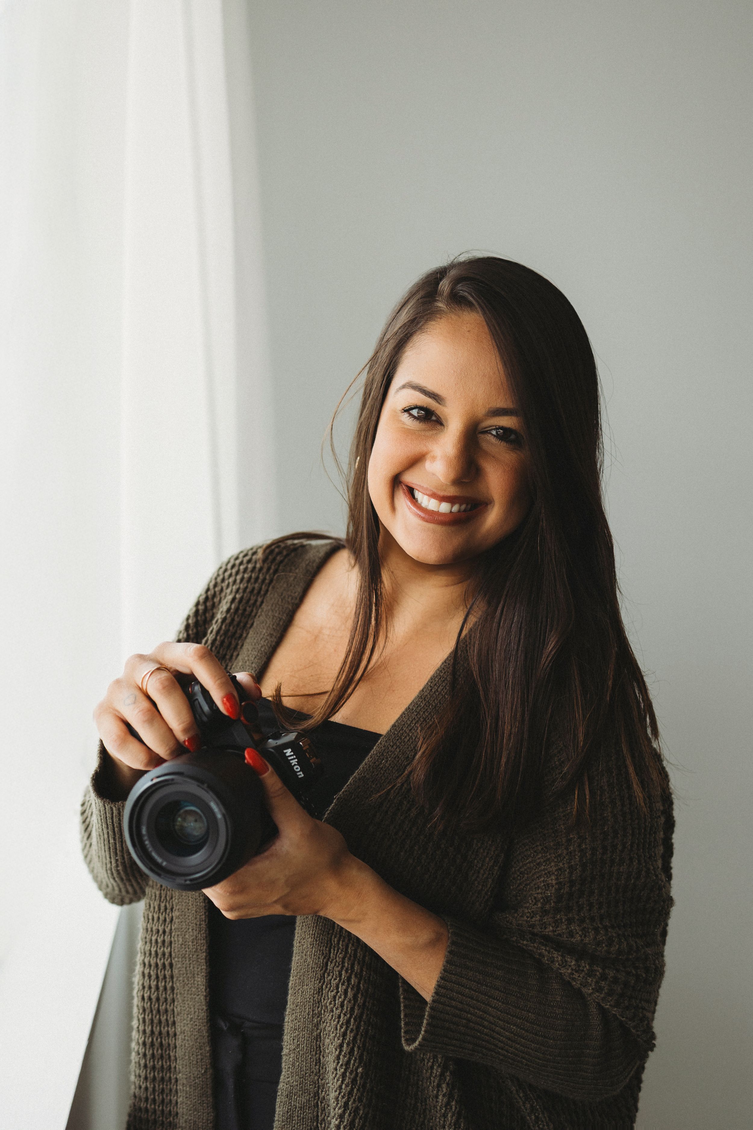 Photographer in brown sweater holding camera against white wall.