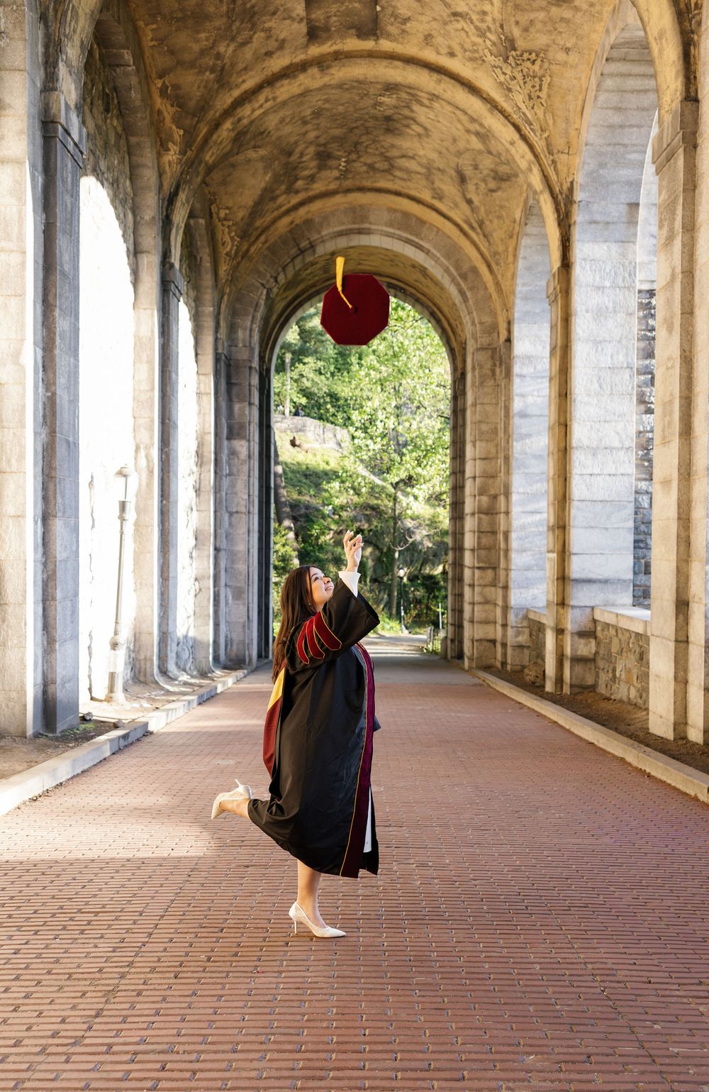Graduate in black gown tosses cap under stone archway colonnade, celebrating academic achievement outdoors.