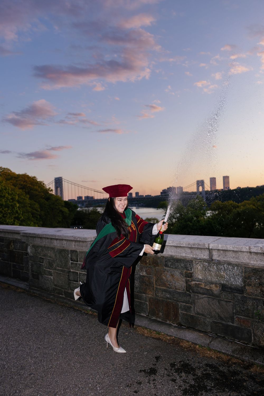 Graduate in cap and gown celebrates with champagne at sunset, George Washington Bridge visible in background.