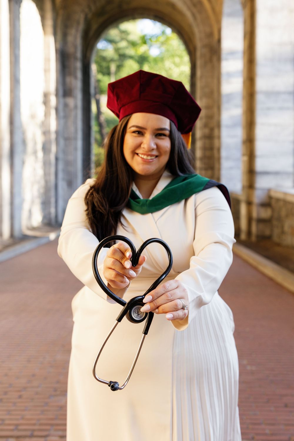 Medical school graduate in white gown and red cap holds stethoscope in heart shape, smiling outdoors.