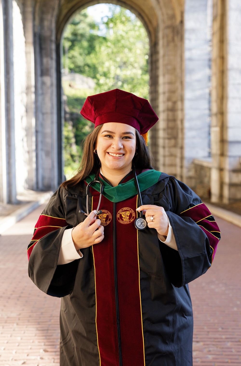 Smiling woman in black doctoral graduation gown with red velvet cap and green hood holding a stethoscope outdoors.