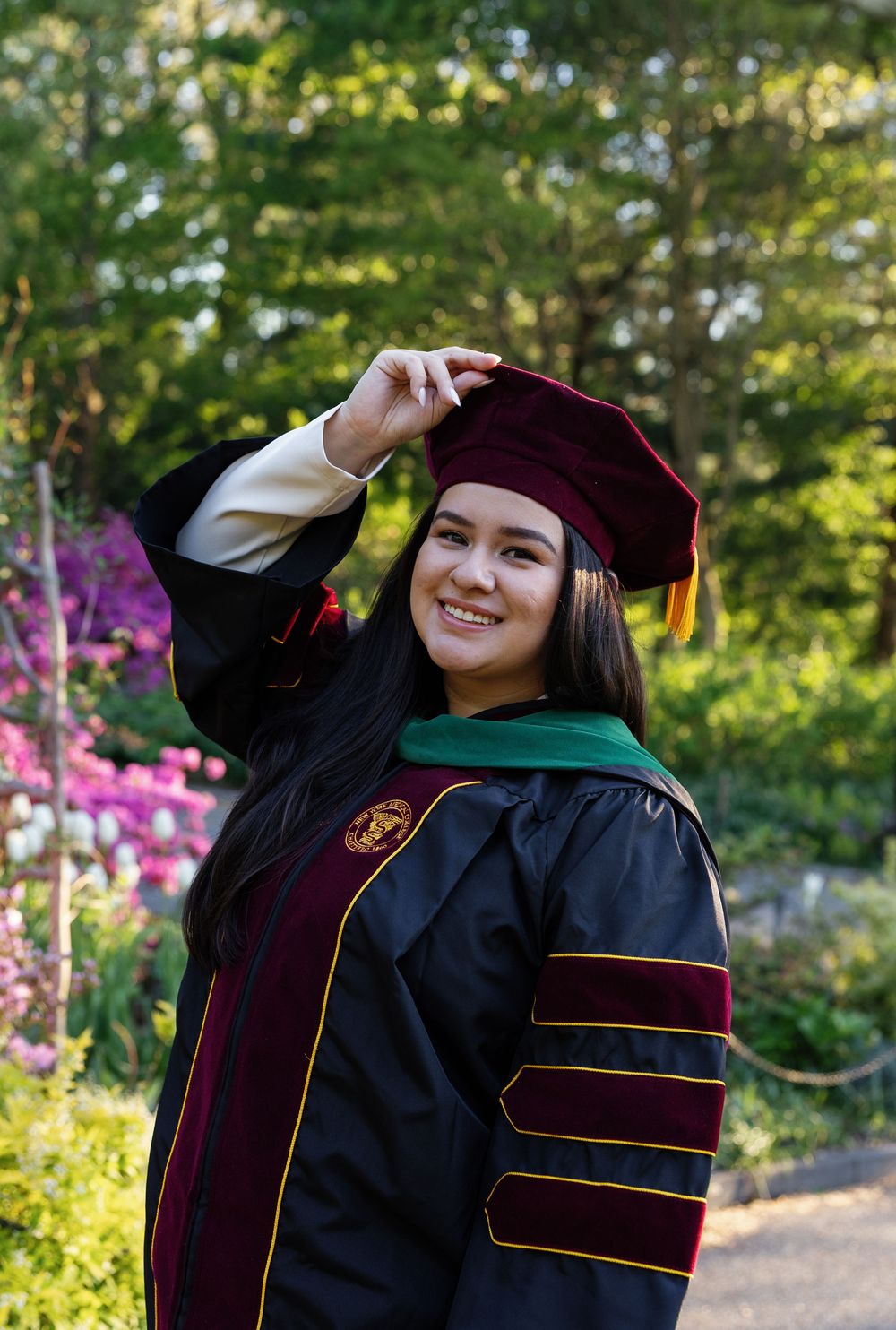 Smiling graduate in black and maroon doctoral regalia holds cap outdoors among colorful flowers and green trees.